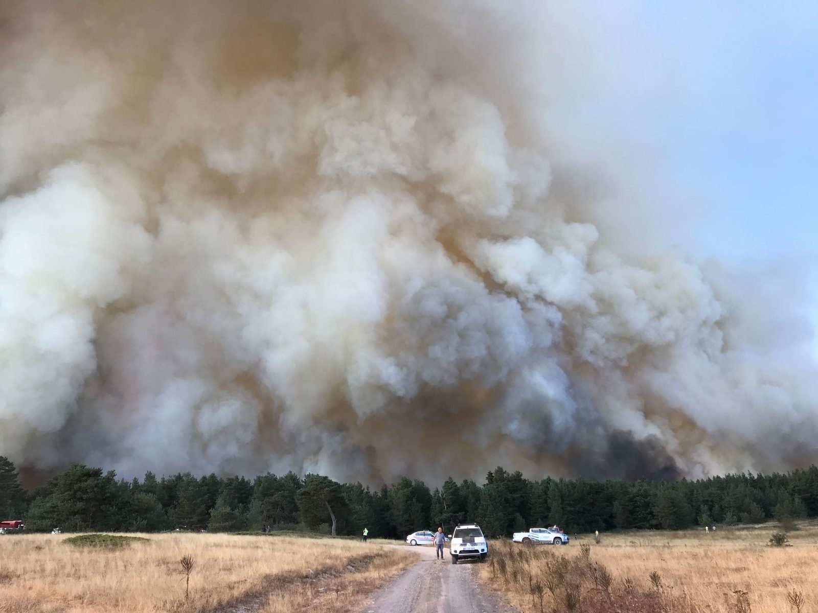 A wildfire smoke column towering over a dirt staging road and parked vehicles