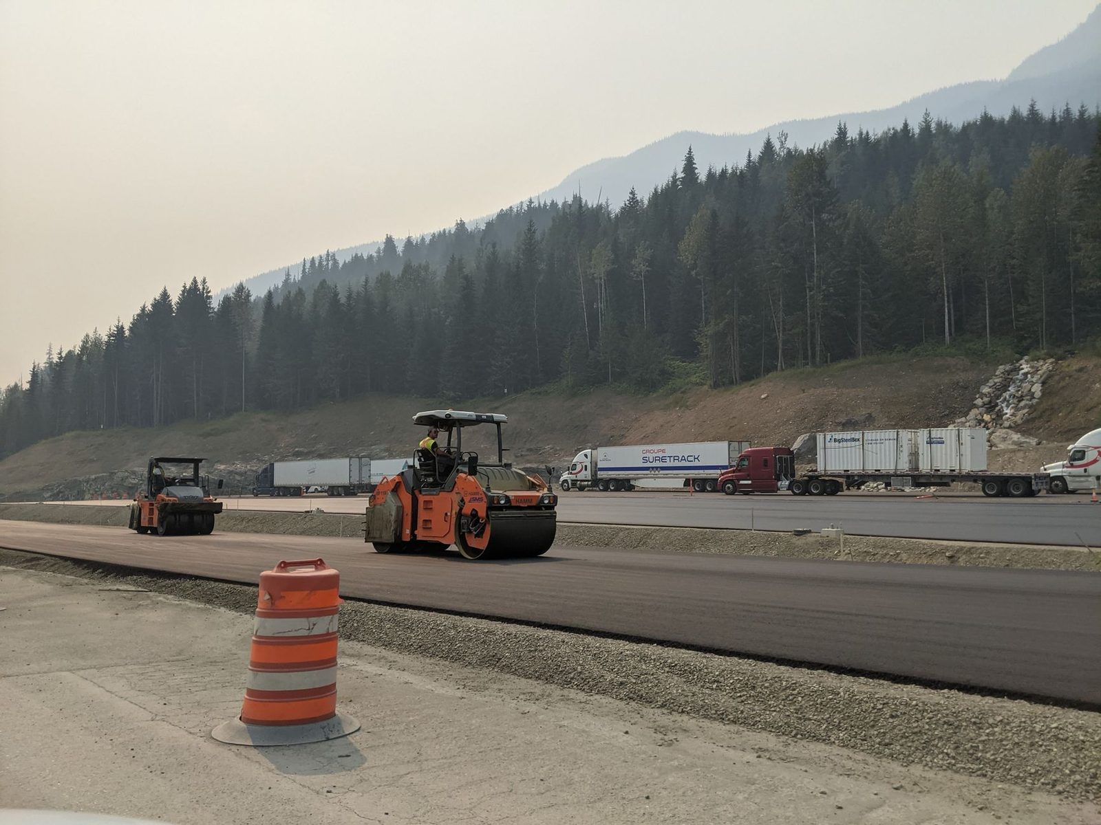 A paving crew rolling fresh asphalt on a new highway lane below forested mountain slopes