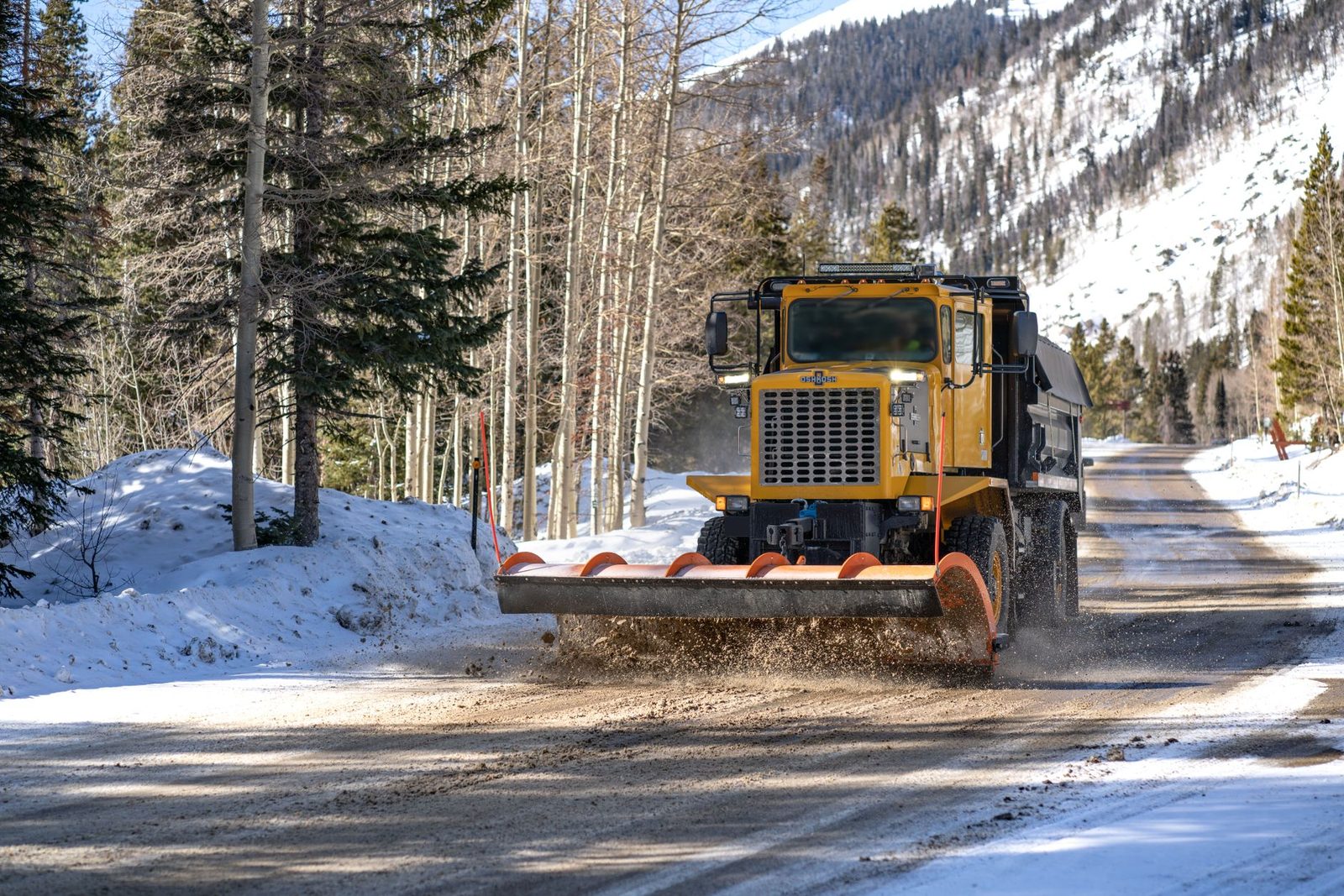 A heavy plow clearing snow from a mountain road through pine and aspen forest